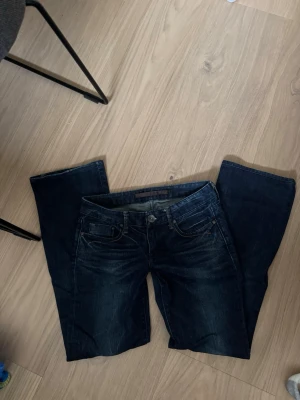 Mörkblå bootcut jeans - Säljer ett par mörkblå bootcut jeans med klassisk femficksdesign och snygga slitningar framtill. Jeansen har kontrastsömmar och stängs med knapp och dragkedja. Perfekta för dig som gillar en tidlös och avslappnad stil. Tappat några stenar på ena fickan ibak! 