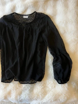 Svart blus - Snygg svart blus i lite genomskinligt material med diskreta prickar och knytband vid halsen. 