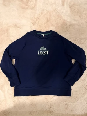 Mörkblå sweatshirt från Lacoste - Mörkblå sweatshirt med rund hals och lång ärm från Lacoste. Tröjan har en broderad logotyp med krokodil och texten 'LACOSTE' på bröstet. Klassisk och stilren design som passar perfekt till jeans eller joggers.