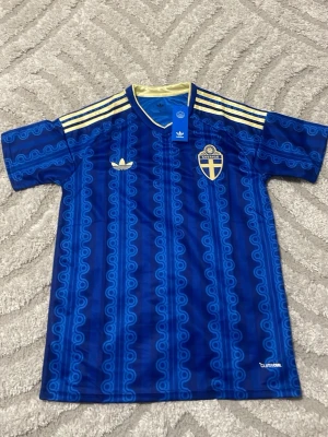 Blå Sverige fotbollströja Adidas - Säljer en blå fotbollströja från Adidas med Sveriges landslagsemblem på bröstet. Tröjan har gula detaljer vid kragen och axlarna samt ett unikt mönster i olika blå nyanser. Korta ärmar och lätt, ventilerande material. Perfekt för match eller träning.