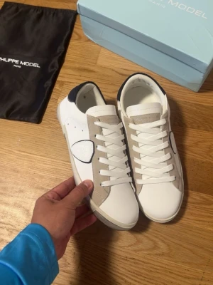 Vita Philippe Model sneakers med beige detaljer - Snygga vita sneakers från Philippe Model med beige mockadetaljer och mörkblå häl. Skorna har klassisk låg profil, vita skosnören och en stilren design med märkets logga på sidan. Perfekta för en trendig och avslappnad look.