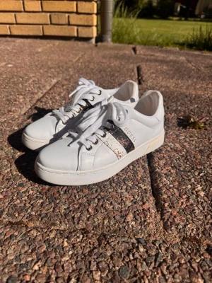 Vita sneakers från Guess - Snygga vita sneakers från Guess med klassisk låg modell. Skorna har vita snören, detaljer i silver och svart samt Guess-logga på sidan. Tillverkade i slätt material med diskret mönster på sidan. Perfekta för en trendig och stilren look.