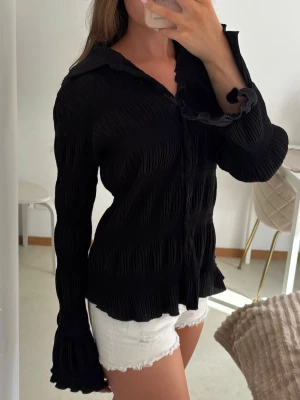 Svart plisserad blus/skjorta  - Superfin långärmad blus/skjorta med plissering och volanger nertill. Lappen är avklippt men skulle uppskatta storlek 36/38