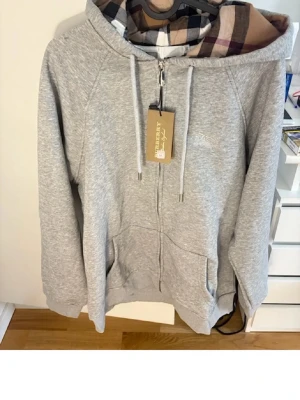 Grå zip hoodie med rutigt foder - Snygg grå hoodie med dragkedja och huva. Insidan av huvan har ett klassiskt rutigt mönster i beige, svart och vinrött. Tröjan har fickor framtill och justerbara snören i huvan. Perfekt för en chill och avslappnad stil.