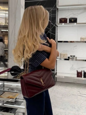 Vinröd YSL handväska i läder - Säljer en vinröd handväska från YSL i quiltat läder med guldfärgad kedjerem och ikonisk YSL-logga framtill. Väskan har en klassisk rektangulär form och elegant design som passar till många olika looks. Perfekt accessoar för dig som gillar lyxiga detaljer.