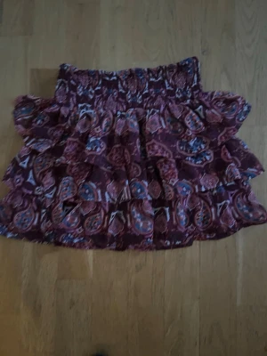 Mönstrad volangkjol från Lindex - Kort volangkjol från Lindex i vinrött med paisleyinspirerat mönster i blått, vitt och rosa. Kjolen har resår i midjan och flera lager volanger som ger en lekfull look. Perfekt för dig som gillar färg och mönster.