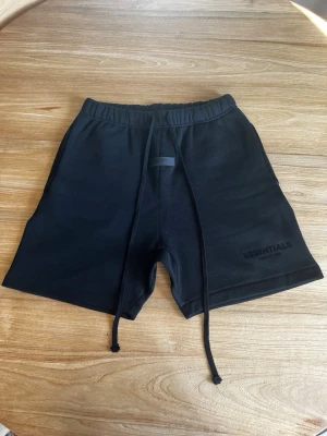 Svarta shorts Essentials Fear of God - Säljer ett par svarta shorts från Essentials Fear of God med långa snören i midjan och diskret logga broderad på benet. De har en relaxed fit och elastisk midja för extra komfort. Perfekta för chill dagar eller streetwear-stil.
