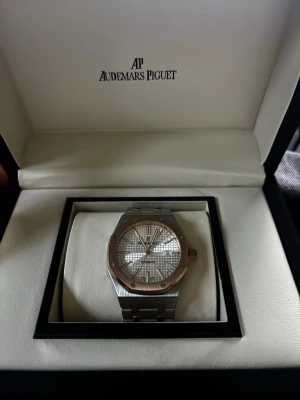 Audemars Piguet Royal Oak klocka - Exklusiv Audemars Piguet Royal Oak klocka i silver med ikonisk åttakantig boett och rutmönstrad urtavla. Klockan har detaljer i guldton och länkarmband i metall. Perfekt för dig som gillar lyxiga accessoarer.