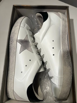 Vita Golden Goose med black tail - Snygga vita sneakers från Golden Goose med grå stjärndetalj på sidan. Skorna är helt nya med snygga slitningar. Skorna är i Storlek 43 men passar även 42. 