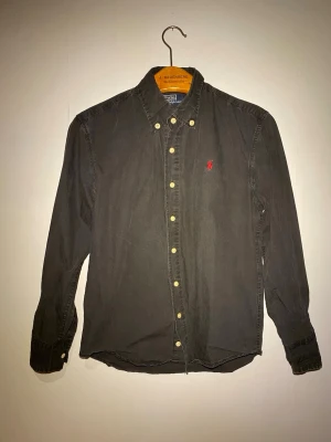 Svart skjorta från Ralph Lauren - Svart skjorta från Polo Ralph Lauren med klassisk button-down krage och guldfärgade knappar. Broderad röd logga på bröstet. Skjortan är långärmad och tillverkad i mjuk bomull, perfekt för en clean och stilren look.