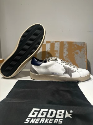 Vita Golden Goose sneakers - Snygga vita sneakers från Golden Goose med grå stjärna på sidan, mörkblå detalj vid hälen, slitningar och beige sula. Skorna är helt nya i storlek 43 men passar även 42. 