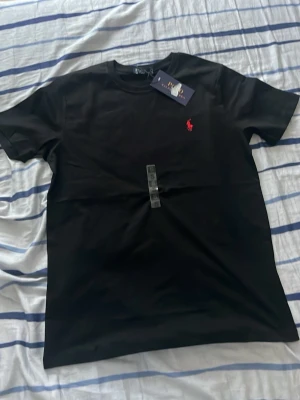 Svart t-shirt från Polo Ralph Lauren - Klassisk svart t-shirt från Polo Ralph Lauren med röd broderad logga på bröstet. T-shirten har rund halsringning och korta ärmar. Tillverkad i mjuk bomull för en skön känsla och enkel stil.
