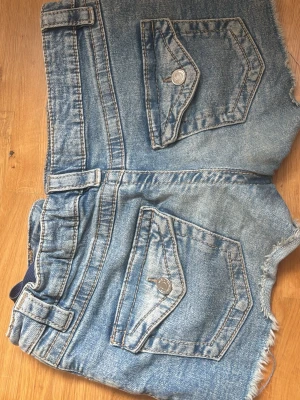 Blå jeansshorts med råa kanter - Säljer ett par blå jeansshorts med råa, fransiga kanter och stora fickor med knappdetaljer. Shortsen har bälteshällor och klassisk femficksdesign. Perfekta för varma dagar och en avslappnad stil.
