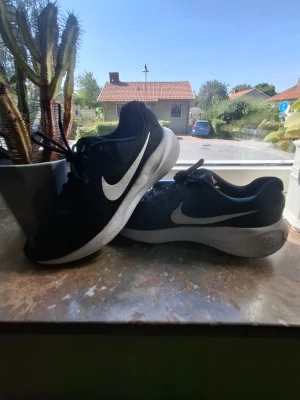 Svarta Nike sneakers med vit sula - Snygga svarta Nike sneakers med vit sula och den klassiska vita swoosh-loggan på sidan. Ovandelen är i meshmaterial som andas och sulan är tjock och bekväm. Perfekta för dig som vill ha en sportig och stilren look. Perfekt för sportande med kompisar medans du fortfarande har ett par snygga skor