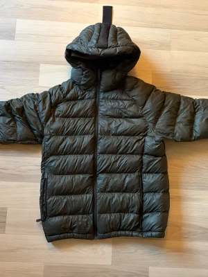  Peak Performance Jacka - Snygg mörkgrön dunjacka från Peak Performance med huva och dragkedja framtill. Jackan har quiltad design och två sidofickor. Perfekt för kalla dagar när du vill hålla värmen och ändå se stilren ut.