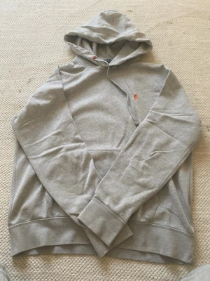 Grå hoodie med huva och broderad detalj - Snygg grå hoodie med huva och dragsko. Tröjan har en liten broderad logga i orange på bröstet och ribbade muddar vid ärmar och nederkant. Perfekt för en avslappnad och stilren look.