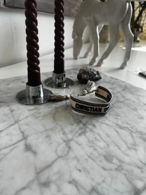 Christian Dior J'Adior armband - Snyggt armband från Christian Dior i beige och svart tyg med texten 'J'ADIOR'. Armbandet har flätad justerbar knytning och dekorativa tofsar i ändarna. Perfekt accessoar för att lyfta din outfit med en exklusiv touch.