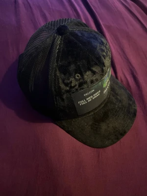 Svart meshkeps med Falcon-tryck - Säljer en svart keps som är limited edition och imte går att köpa någon annan stans. Kepsen har en logga från Falcon Hoppfull ölen tillsammans med ett citat om ölen. Kepsen har böjd skärm och justerbar baksida. Materialet är en mix av tyg och mesh, vilket ger en luftig känsla. Perfekt för dig som gillar streetwear och vill sticka ut. 