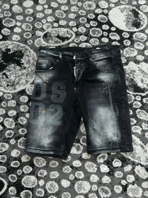Svarta jeansshorts med tryck och ICON-detalj - Säljer ett par svarta jeansshorts med slitningar, tryckta siffror på framsidan och en gul ICON-lapp bak i midjan. Shortsen har fem fickor och dragkedja. Snygg tvättad look och lite längre modell som passar perfekt till sommaren.