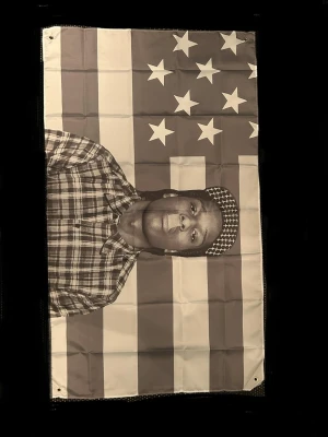 Asap Rocky flag - Säljer en flagga i tyg med ett svartvitt porträtt av en person mot en bakgrund av amerikanska flaggans stjärnor och ränder. Flaggan har öljetter i hörnen för enkel upphängning och är rektangulär i formen.                                                               Size📏: 90cm x 150cm