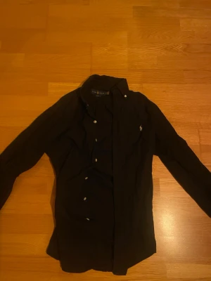 Svart skjorta från Ralph Lauren - Klassisk svart skjorta från Ralph Lauren i slim fit-modell. Skjortan har långa ärmar, knappar framtill och den ikoniska loggan broderad på bröstet. Perfekt för dig som gillar stilrena och tidlösa plagg.