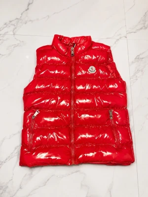 Röd glansig dunväst från Moncler - Snygg röd dunväst från Moncler med glansig finish och två dragkedjefickor framtill. Västen har hög krage och klassisk Moncler-logga på bröstet. Perfekt för lager-på-lager och ger en cool streetstyle-vibe. Priset är ej hugget i sten så kom med bud🤝