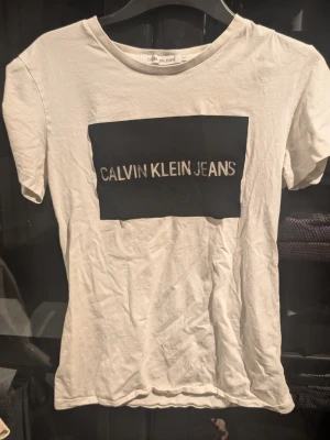 Vit Calvin Klein Jeans t-shirt - Vit t-shirt från Calvin Klein Jeans med klassisk svart logga framtill. T-shirten har rund halsringning och korta ärmar. Tillverkad i mjuk bomull för en bekväm känsla. Inga synliga fläckar eller defekter. Skapa en bundle för bättre pris
