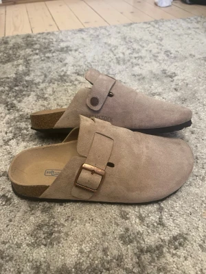 Beige Birkenstock tofflor mocka - Säljer ett par klassiska beige tofflor från Birkenstock i mjuk mocka med justerbart spänne i metall och korksula. Modellen har öppen häl och ergonomisk fotbädd för extra komfort. Perfekta för chill dagar hemma eller när du vill ha det bekvämt.