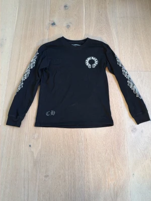 Svart långärmad topp Chrome Hearts - Svart långärmad topp från Chrome Hearts med vita tryck på ärmarna och logga på bröstet. Stort Chrome Hearts-tryck på ryggen och coola detaljer längs båda ärmarna. Tillverkad i mjuk bomull och har en normal passform.