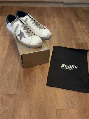 Vita GGDB sneakers med grå stjärna och blå sula - Snygga vita GGDB sneakers med grå stjärndetalj på sidan och gråa skosnören. Skorna har en låg silhuett och mörkblå häl. Tillverkade i läder med slitstark gummisula. Perfekta för en trendig och avslappnad look.
