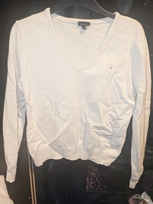 Vit stickad tröja från Gant - Klassisk vit stickad tröja från Gant med V-ringning och diskret logga broderad på bröstet. Tröjan har långa ärmar och ribbade muddar vid ärmslut och nederkant. Inga synliga fläckar eller defekter. Skapa en bundle för bättre pris