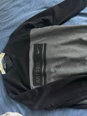 Nike svart och grå sweatshirt - Nike sweatshirt i svart och grått med rund hals och texten 'JUST DO IT.' i relief på bröstet. Tröjan har långa ärmar och en sportig design med mesh-detalj över texten. Perfekt för en avslappnad och trendig look. OBS! storlek S/M Inte xl!!