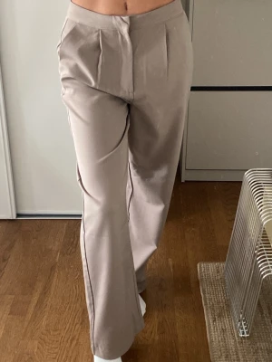 Beige vida kostymbyxor - Säljer ett par beige kostymbyxor med hög midja och vida ben. Byxorna har pressveck framtill och sidofickor. Använda 1 gång.