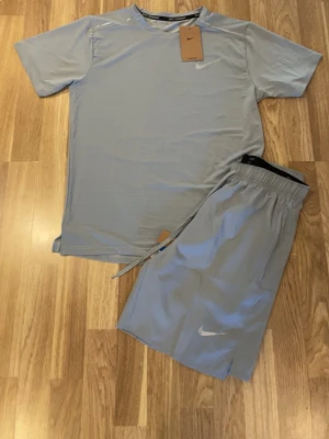Grått träningsset från Nike - Snyggt grått träningsset från Nike med t-shirt och shorts. T-shirten har rund hals och korta ärmar, medan shortsen har resår i midjan och dragsko för bästa passform. Båda plaggen har Nike-logga framtill. Perfekt för gymmet eller löprundan.