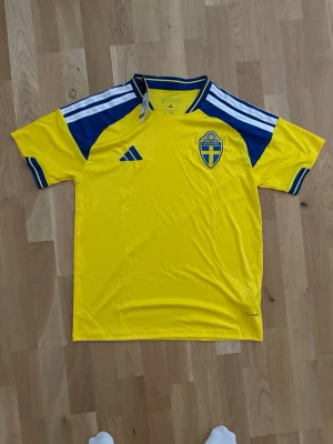 Svensk landslagströja Adidas gul/blå - Gul svensk landslagströja från Adidas med blå detaljer och vita ränder på axlarna. Tröjan har Sveriges landslagsemblem på bröstet och klassisk Adidas-logga. Perfekt för fotboll och sportiga aktiviteter.