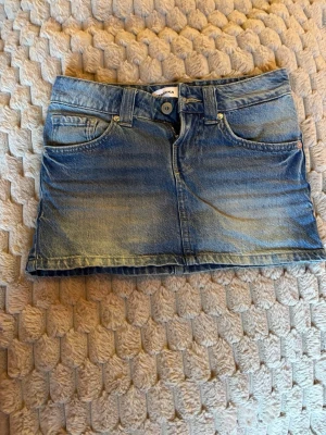 Blå jeanskjol i kort modell - Säljer en klassisk blå jeanskjol. Kjolen har en snygg tvättad look och stängs med knapp och dragkedja framtill. Perfekt till sneakers eller boots för en avslappnad stil.