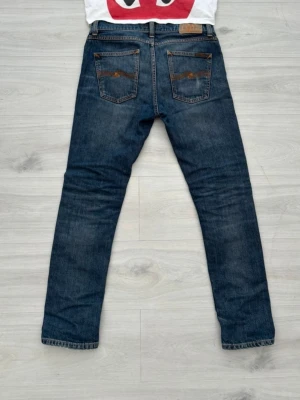 Nudie jeans - Säljer ett par blå jeans med smal passform och klassisk femficksdesign. Jeansen har kontrastsömmar och en mörk tvätt med lätt slitning på framsidan. Perfekta för dig som gillar en enkel och tidlös stil.