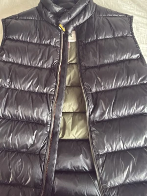 Svart dunväst med hög krage - Snygg svart dunväst med hög krage och dragkedja framtill. Västen har breda, quiltade paneler och en olivgrön insida. Perfekt för lager-på-lager och håller dig varm under kyliga dagar. Passar till många olika stilar. STORLEK M FÖR BAR, så ungefär S-XS för vuxna 