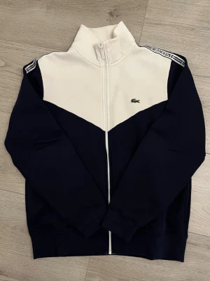 Ziptröja Lacoste - Snygg ziptröja från Lacoste i marinblått och vitt med hög krage och dragkedja framtill. Tröjan har ikoniska Lacoste-loggan på bröstet och vita detaljer/lacoste-band på axlarna och armarna. 