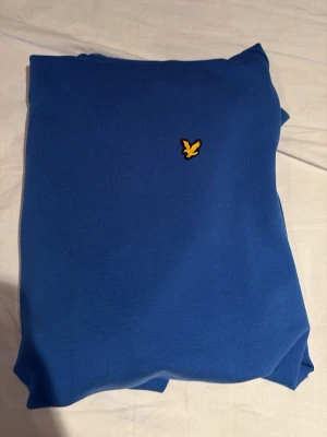 Blå hoodie med gul logga - Snygg blå hoodie med huva och magficka. På bröstet sitter en liten gul broderad logga i form av en fågel. Hoodien har en klassisk passform och är gjord i mjukt bomullsmaterial. Perfekt för en chill och avslappnad stil.