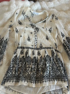 Vit och svart kortklänning från Zara - Supersöt kortklänning från Zara i vit bomull med svart broderat mönster. Klänningen har långa ärmar, v-ringning med snörning och volang nedtill. Perfekt för sommarens alla tillfällen och ger en bohemisk vibe.