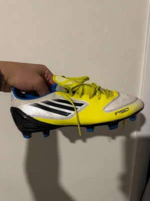 Adidas F50 gula fotbollsskor - Säljer ett par Adidas F50 fotbollsskor i en riktigt snygg gul och vit färgkombination med svarta detaljer och blå dobbar. Skorna har klassisk snörning och är tillverkade i syntetmaterial för lätt känsla på planen. Perfekta för dig som vill sticka ut på fotbollsplanen!