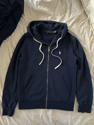 Mörkblå hoodie från  Ralph Lauren (dm för pris - Snygg mörkblå hoodie med dragkedja från Polo Ralph Lauren. Klassisk design med vit logga på bröstet, vita snören i huvan och två fickor framtill. Perfekt för en avslappnad och stilren look.