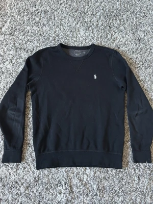 Svart sweatshirt Polo Ralph Lauren - Klassisk svart sweatshirt från Polo Ralph Lauren med rund halsringning och diskret broderad logga på bröstet. Tröjan har långa ärmar och ribbade muddar vid ärmslut och nederkant. Perfekt för en stilren och avslappnad look. Det är en liten fläck med målarfärg dock.