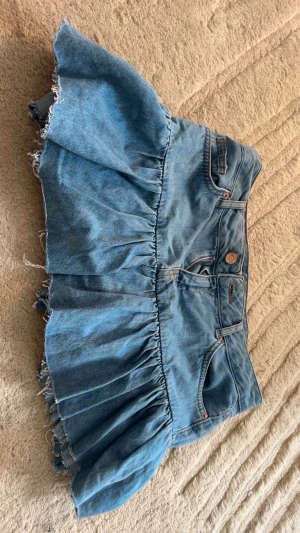 Kort jeanskjol med volang - Säljer en blå kort jeanskjol med volangkant och råa, fransiga kanter. Kjolen har klassiska jeansdetaljer som fickor fram och bak samt knapp och dragkedja i midjan. Perfekt för dig som gillar en avslappnad och trendig stil.