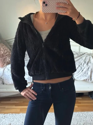 Svart teddyfleece med dragkedja - Mysig svart fleece med dragkedja framtill och huva. Fleecen har en croppad passform och ribbade muddar vid ärmslut och nederkant. Perfekt för att hålla värmen och samtidigt se snygg ut. Kan användas som en fleece ute men även inne som en tröja!💕