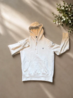 Vit hoodie från Polo Ralph Lauren - Säljer en stilren vit hoodie från Polo Ralph Lauren med klassisk huva och dragsko. Tröjan har en känguruficka framtill och den ikoniska lilla loggan broderad på bröstet. Perfekt för en avslappnad och trendig look. Tröjan har två jättesmå blåa prickar på högra ärmen (inget som syns), skicka iväg ett pm för bild på det. Inga defekter förutom det och att den är lite noppig (syns inte heller)