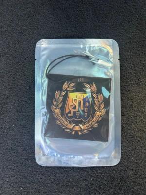 Bildoft SSK - En svart doftgran med Sportklubbens emblem i guld och blått, omgiven av en lagerkrans. Produkten har ett snöre för upphängning och levereras i försluten plastförpackning. Perfekt för att fräscha upp bilen eller rummet.