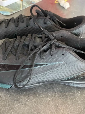 Svarta fotbollsskor med snörning - Säljer ett par svarta fotbollsskor med snörning och låg profil. Skorna har en diskret design med tunna linjer i turkos längs sidan och är tillverkade i syntetmaterial för bra grepp och känsla på planen. Perfekta för dig som vill ha stilrena och funktionella skor till matchen.