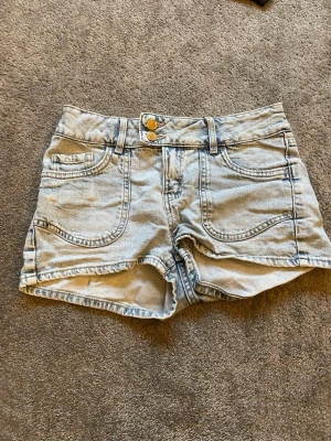 Ljusblå denimshorts med broderade fickor - Säljer ett par ljusblå denimshorts med dubbla knappar och broderade bakfickor i hjärtformad design.  Perfekta för varma dagar!